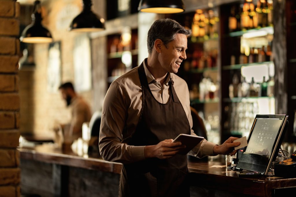 Male waiter using EPOS system on counter showing EPOS features Ireland essential vs premium POS system functionality